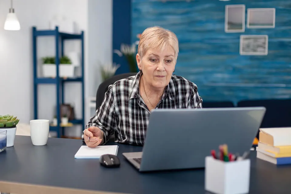 A focused older woman working on a laptop with a notebook and pen on her desk.