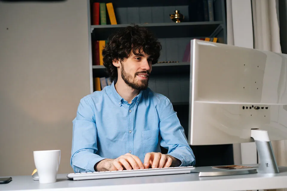 A smiling man with curly hair working at a desktop computer in a home office setting.