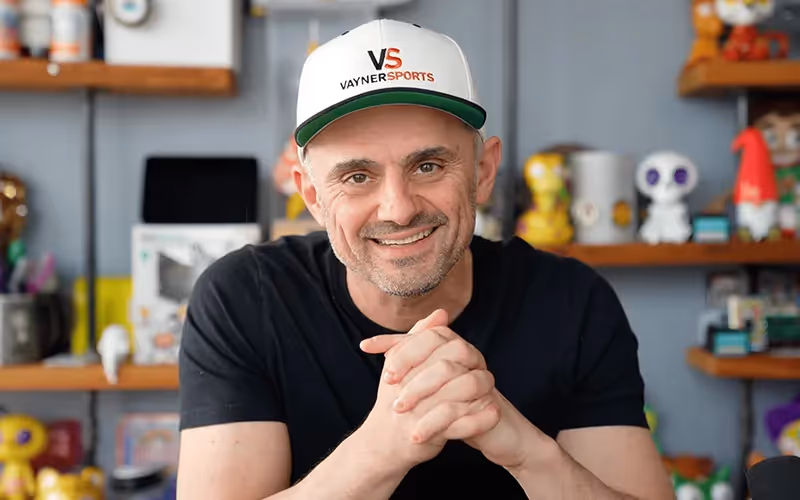 Gary Vaynerchuk, a white man wearing a cap is sitting down and smiling towards the camera. Behind him are shelves of figurines.