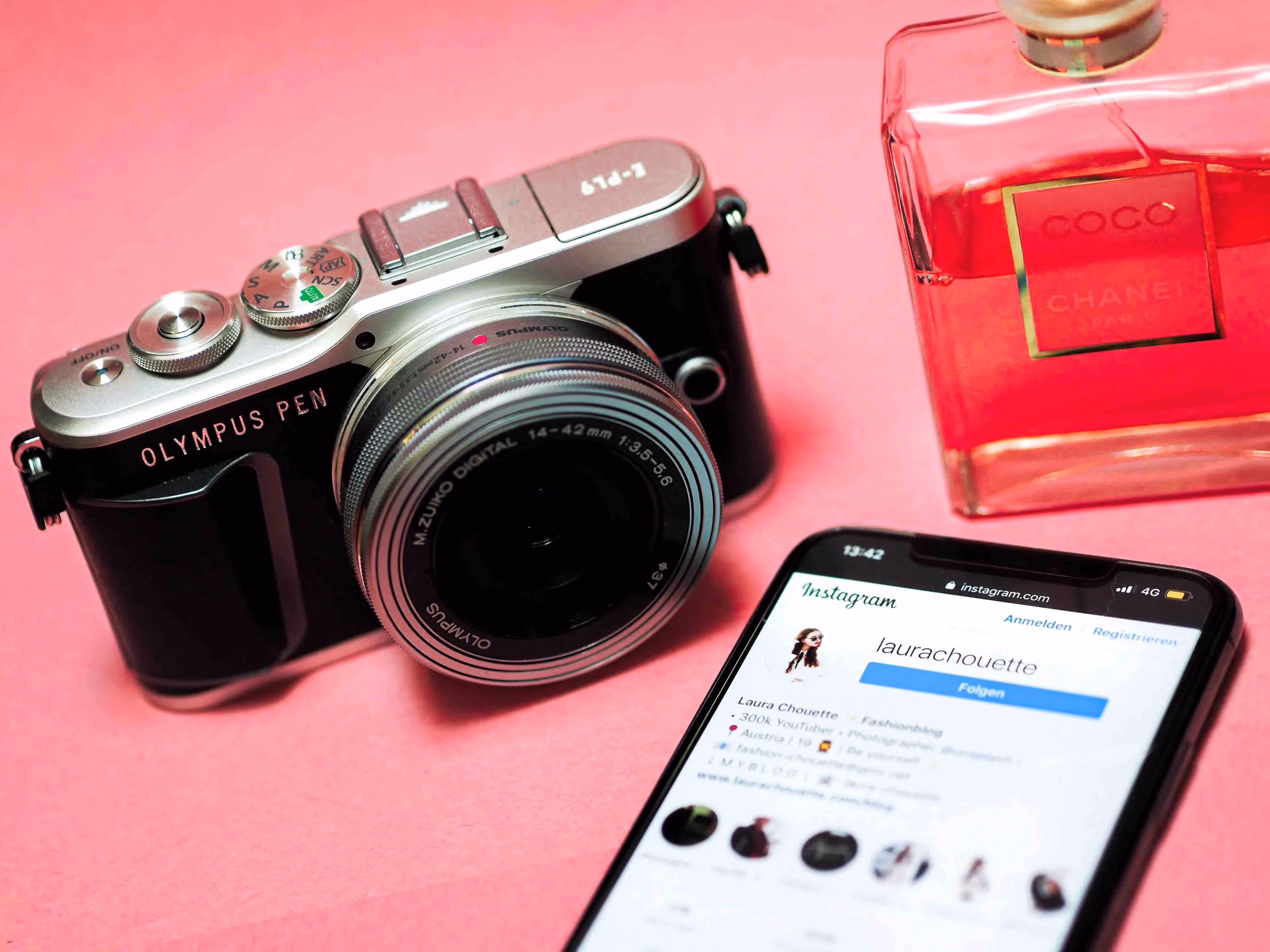A camera, iPhone, and perfume bottle on a pink table.