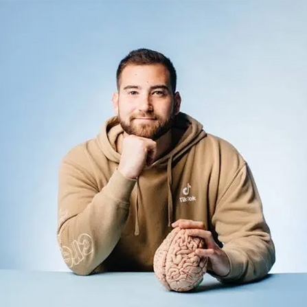 Image shows Neuoscientist Ben Rein, a young white man sitting at a desk holding a model brain.