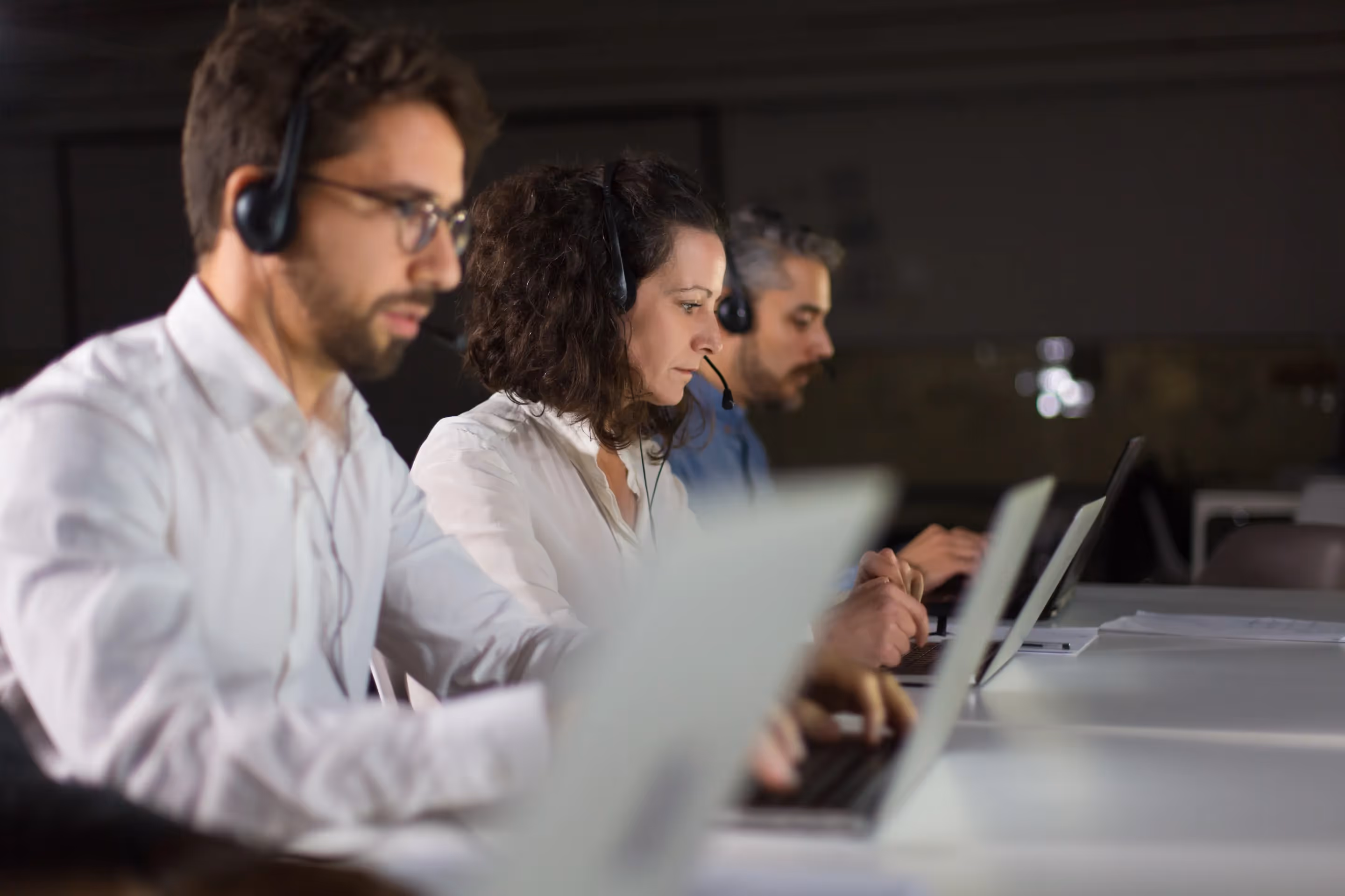 Three Customer services employees wearing headsets, engaged in conversation while seated at their desks.