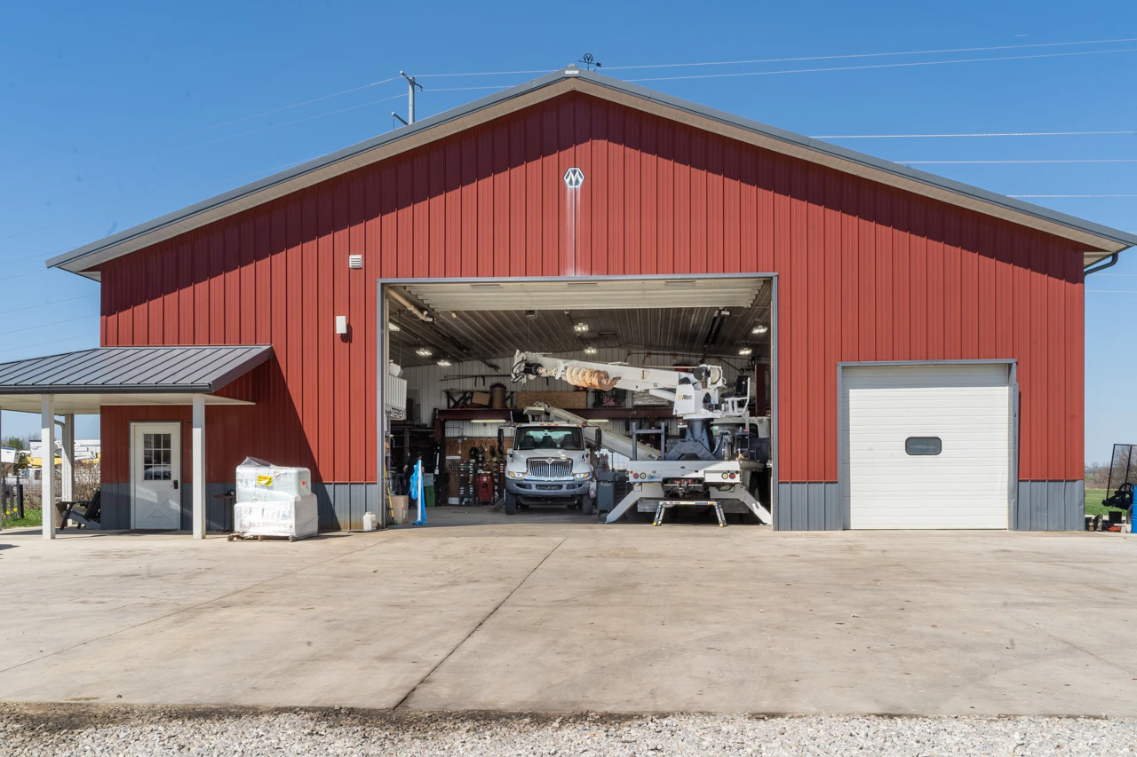 A large red metal workshop building with a gray roof and white trim stands under a clear blue sky, its garage door open to reveal a white utility truck with a mounted crane inside. The concrete driveway in front is clean, with equipment neatly arranged.