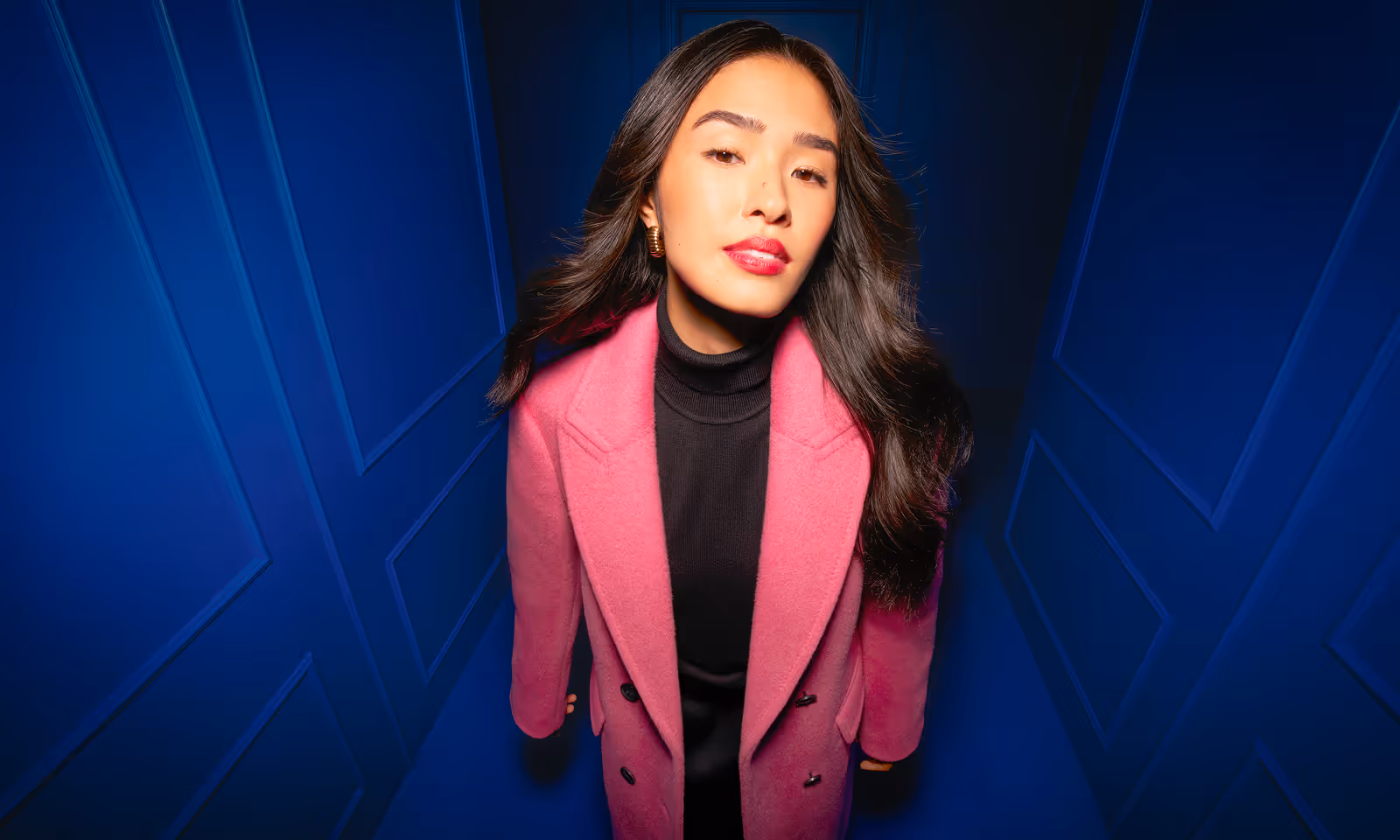 RW&Co Holiday 2025 Campaign model with long dark hair wearing a pink coat and black turtleneck standing against a blue paneled wall.