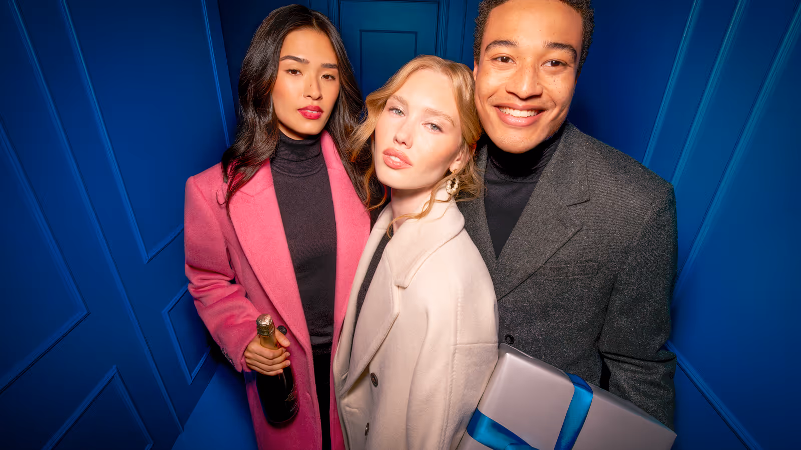 RW&Co Holiday 2025 Campaign three friends dressed in winter coats, one holding a champagne bottle and another a wrapped gift with blue ribbon, posing against a blue wall.