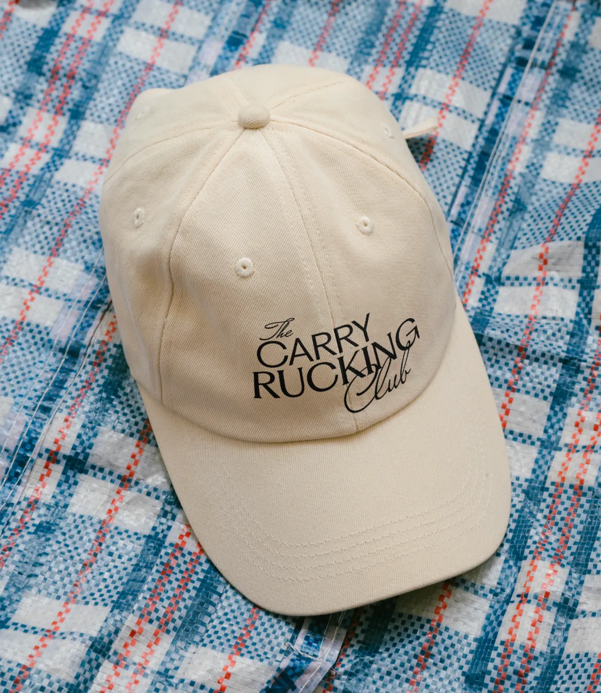 Beige baseball cap with black text reading 'The Carry Rucking Club' placed on a blue, white, and red plaid fabric background.
