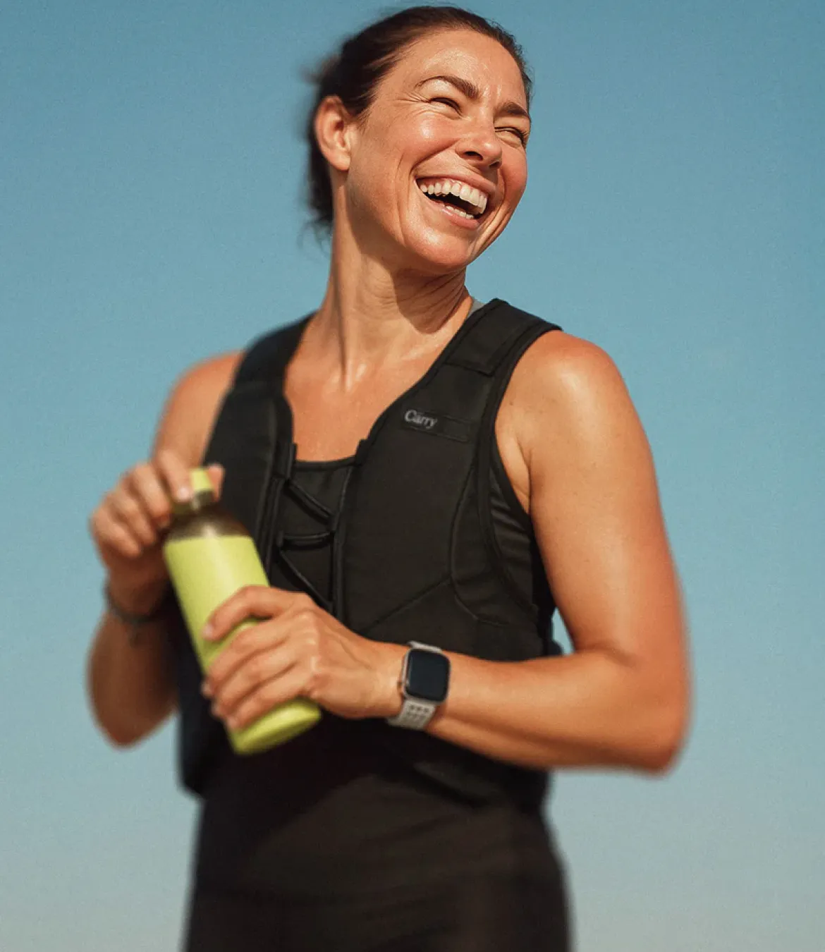 Smiling athletic woman wearing a black weighted vest and smartwatch, holding a green water bottle against a clear blue sky.