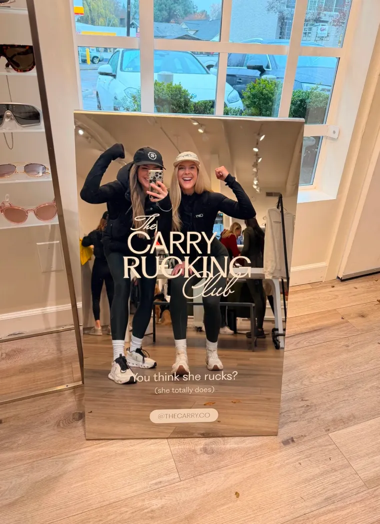 Two women dressed in sporty black outfits and caps flexing arms and smiling in a store mirror with The Carry Rucking Club logo overlay.