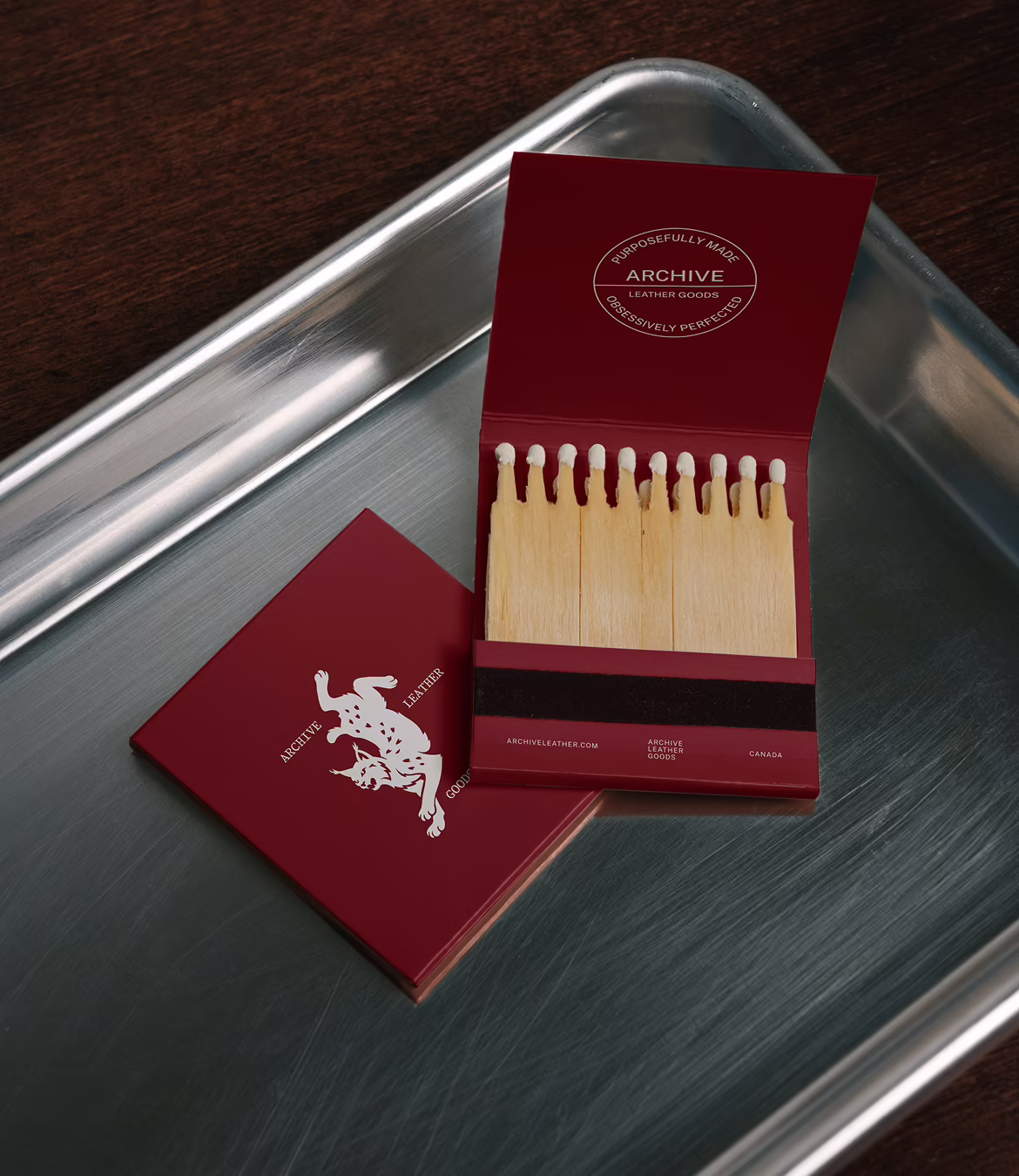 Two red matchbook-style boxes with white text and a frog logo sit open on a stainless steel tray over a dark wooden surface; one box is open showing wooden matches inside.