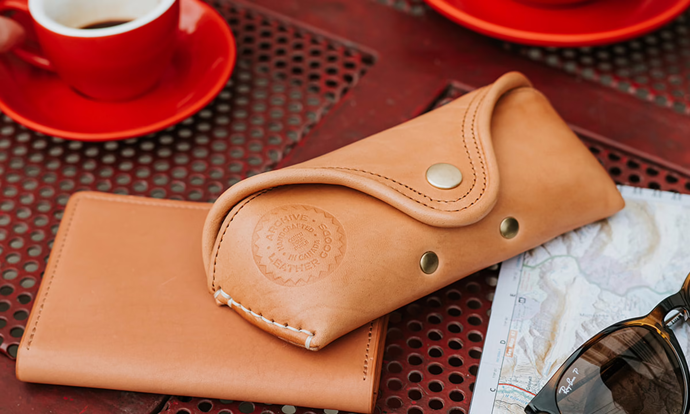 Tan leather sunglasses case and matching wallet placed on a perforated metal table next to a folded map and Ray-Ban sunglasses, with a red cup and saucer of coffee in the background.