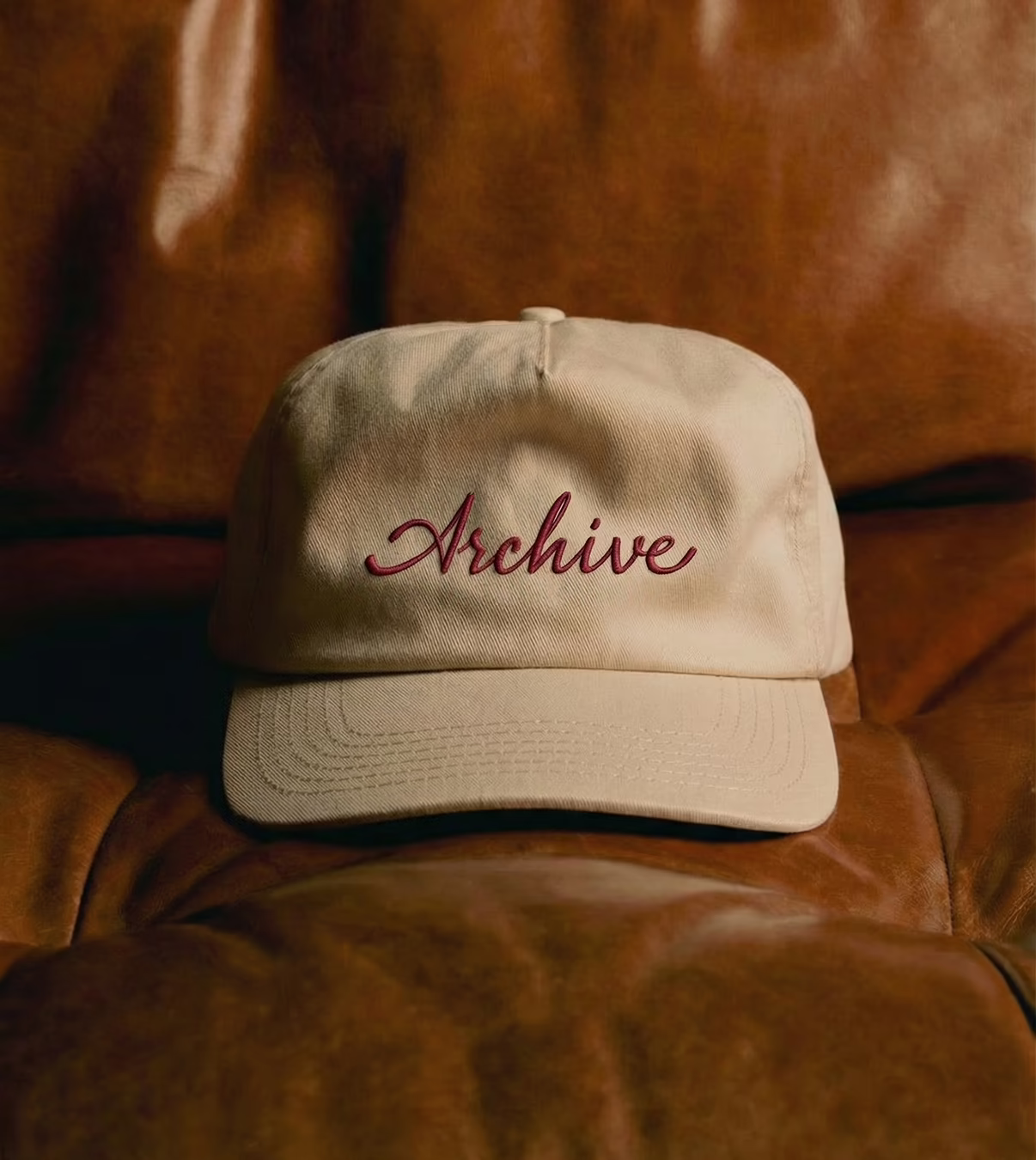 Beige baseball cap with the word 'Archive' embroidered in red script on the front, resting on a brown leather surface.