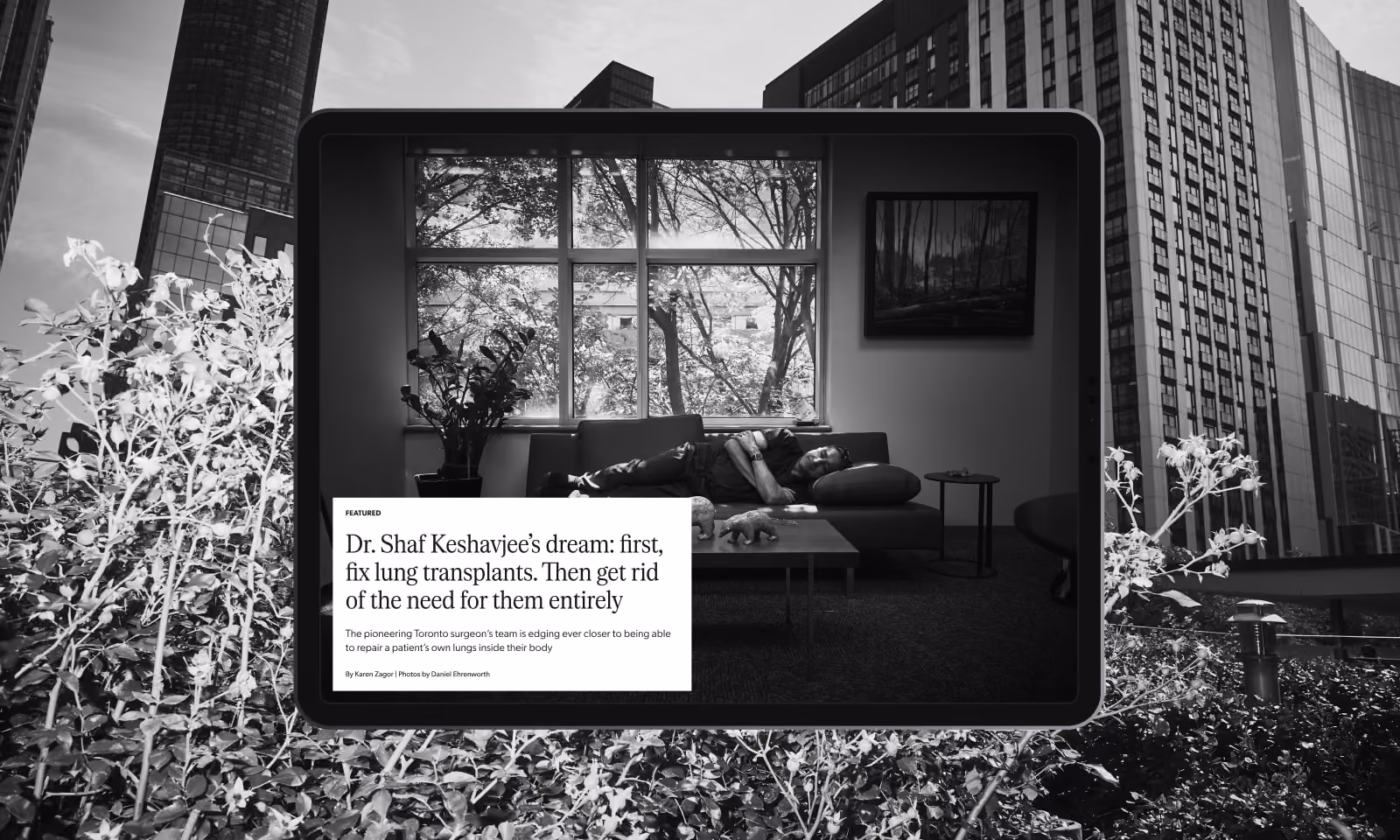 Black and white photograph of a tablet showing a man lying on a couch in a living room with a large window and plants, placed outdoors in front of tall city buildings and flowers.