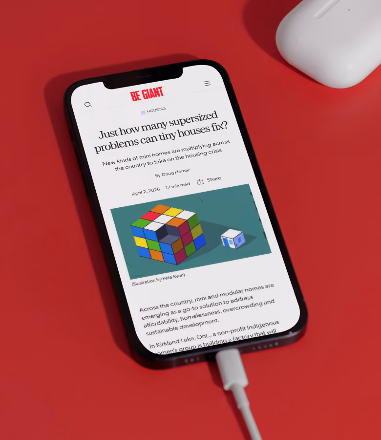 Smartphone on a red surface displaying an article from BE GIANT titled 'Just how many supersized problems can tiny houses fix?' with a colorful Rubik's Cube illustration below the headline.