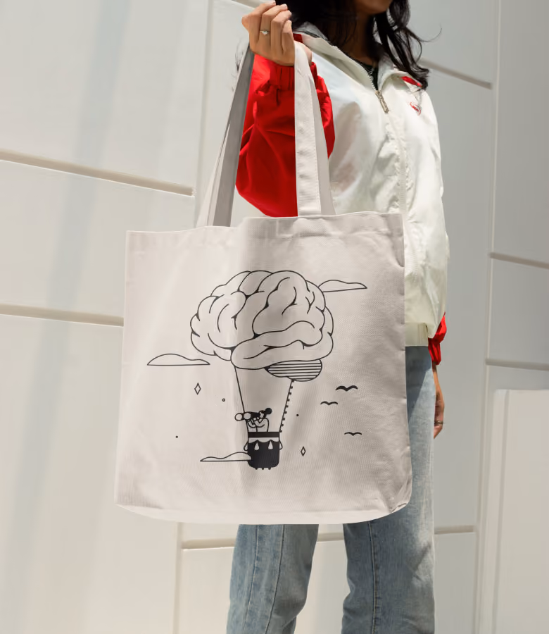 Person wearing a white and red jacket holding a beige canvas tote bag with a black line drawing of a hot air balloon shaped like a brain with birds and clouds around it.
