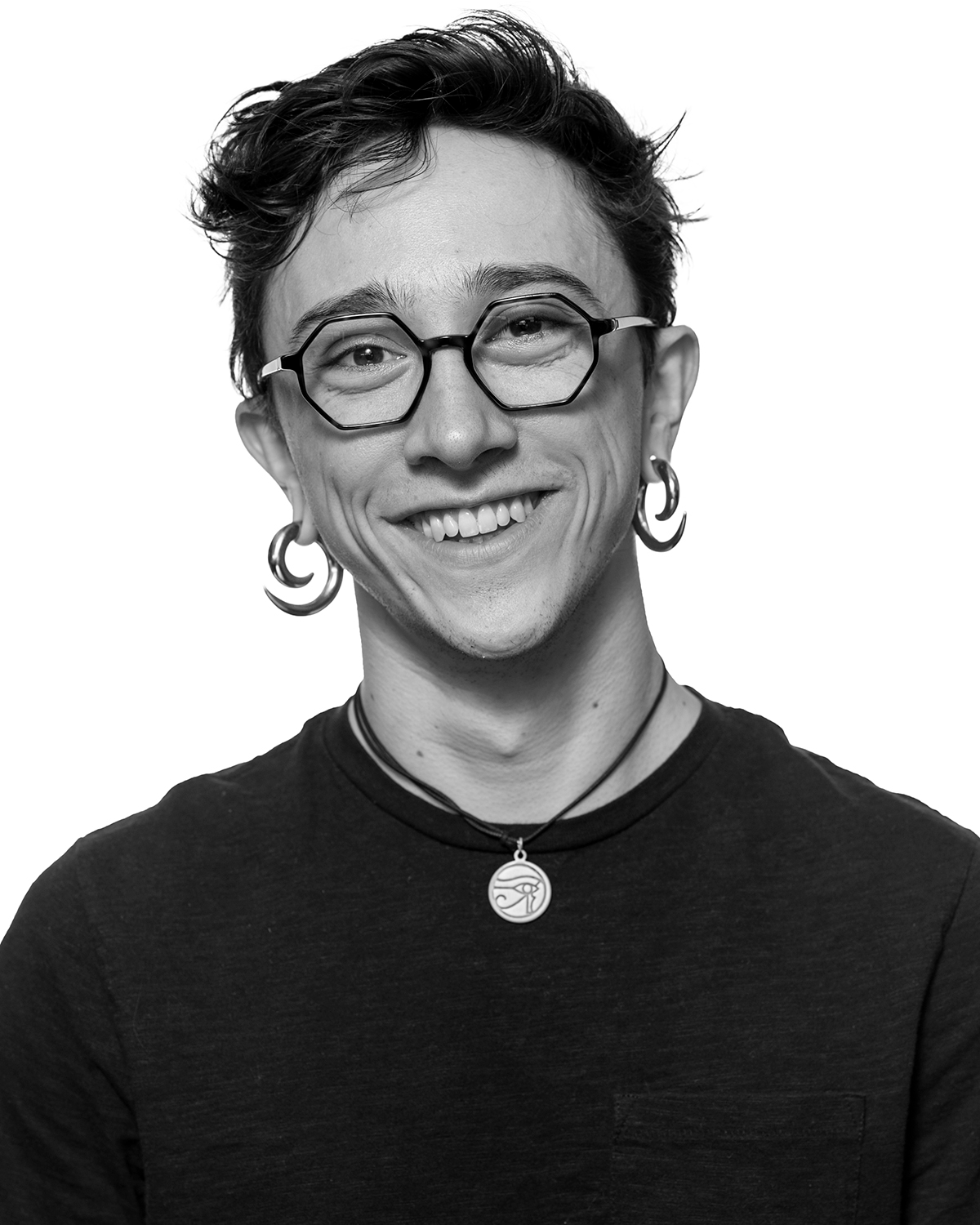 A black and white photo of Django. They are Caucasian, wearing seven sided glasses, silver spiral earrings, an eye of ra necklace, and a black tshirt. They are smiling. 