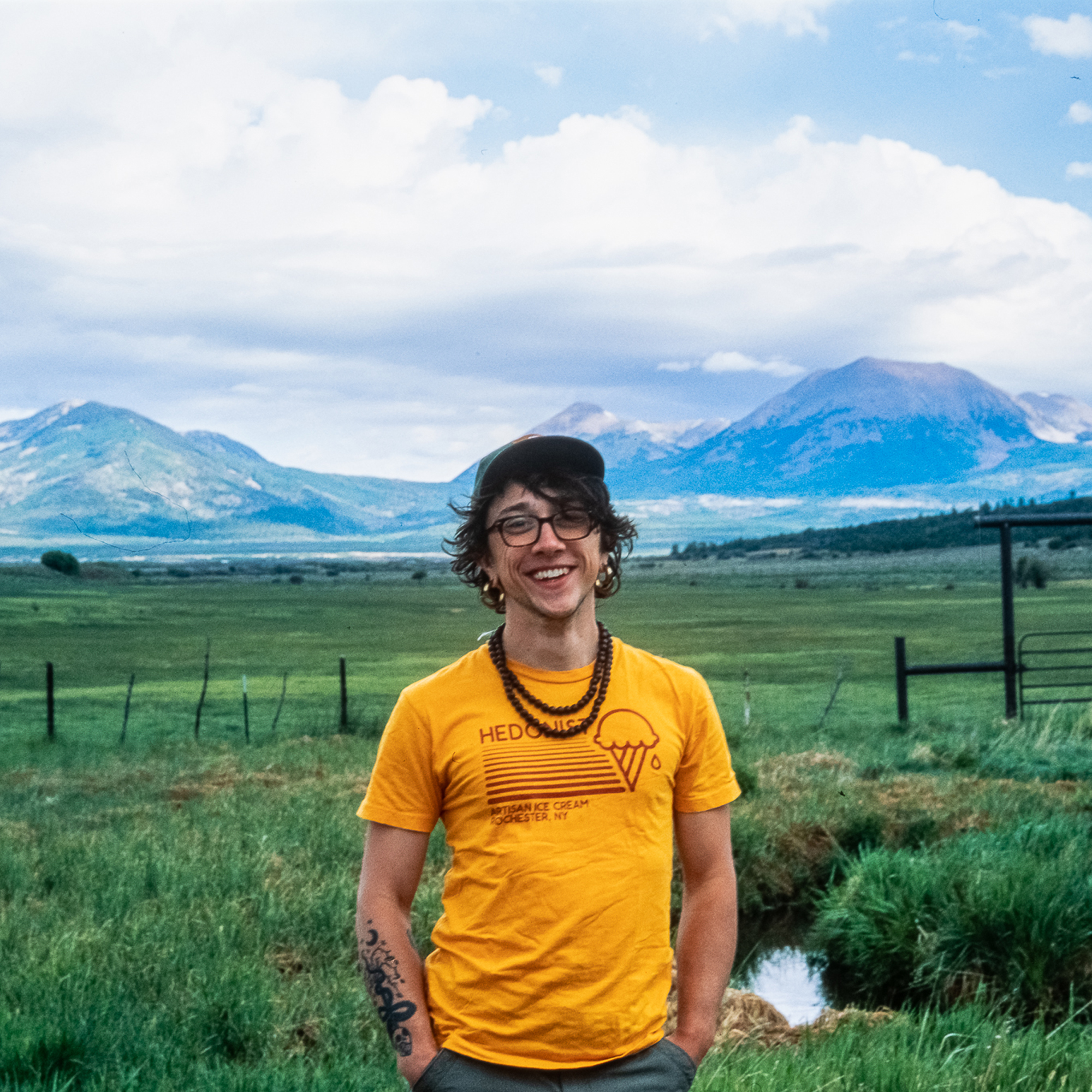 A photo of Django standing at the edge of a field, grasslands stretching for miles behind them to a mountain range in the background. They're wearing a yellow shirt, national park service baseball cap, wayfarer glasses, and mala beads.