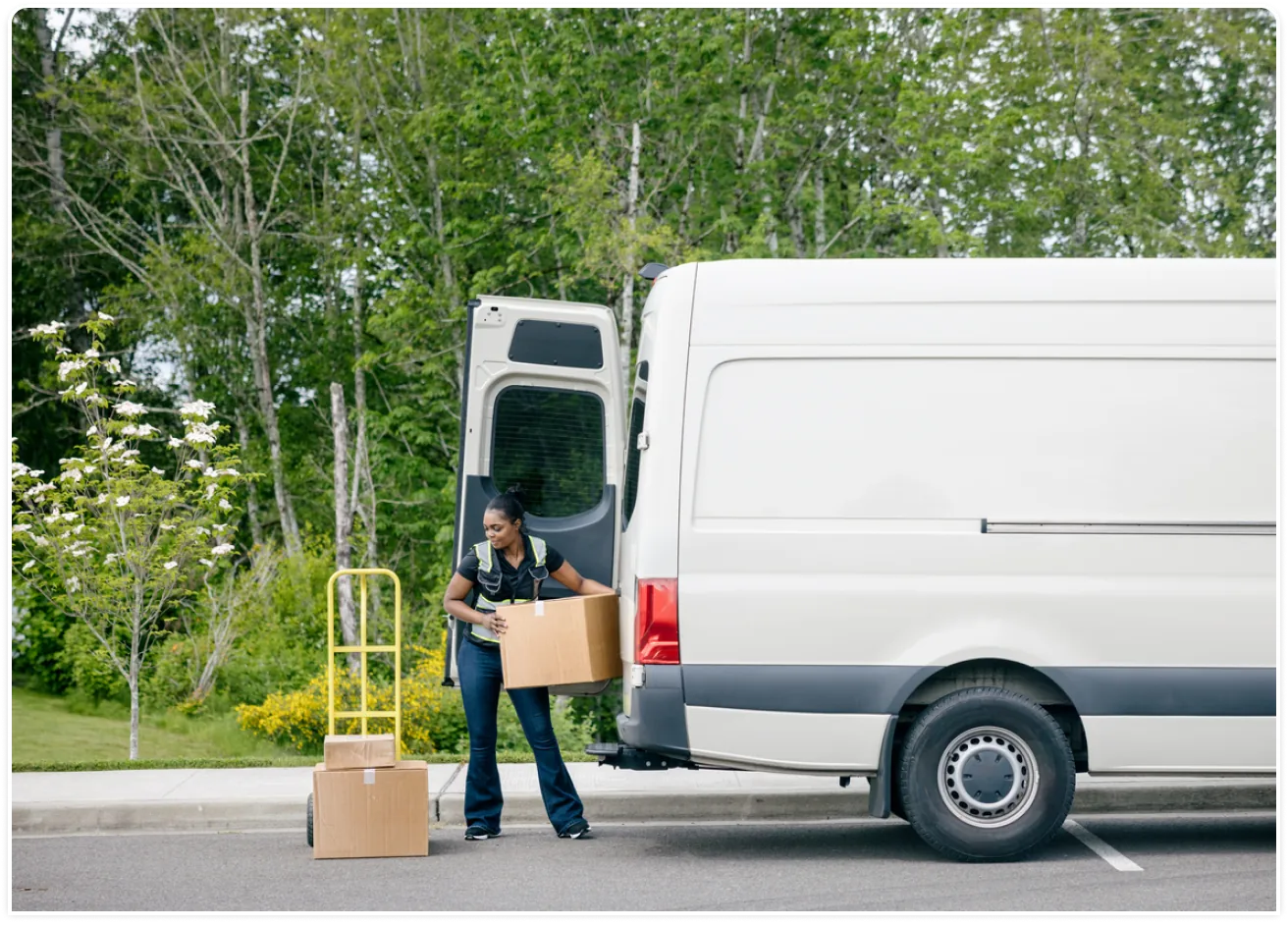 A courier driver unloads boxes from the back of her van onto a hand truck.