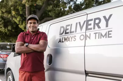 A dark-skinned man dressed in a red polo shirt and trousers stands beside a white van with the words “Delivery. Always on time” written on the side. His arms are crossed and he is smiling slightly. Image credit: RODNAE Productions