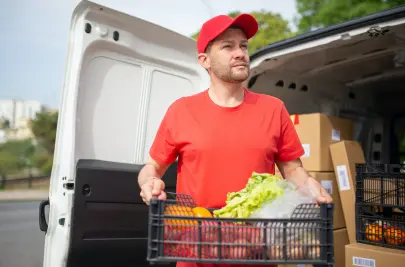 A delivery driver stands by the open door of his van, holding a plastic crate full of fruits and vegetables.