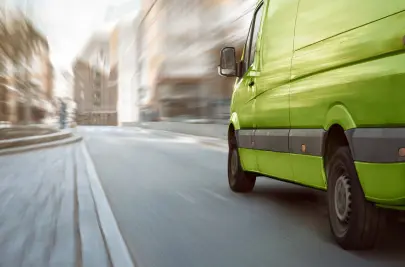 A green delivery van heads away from the viewer down an urban street. 