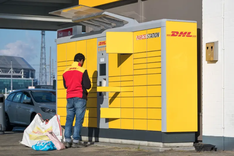 A man in a DHL branded jacket stands at a DHL Parcel Station. There are bags full of packages on the ground, and several delivery locker doors are open. 