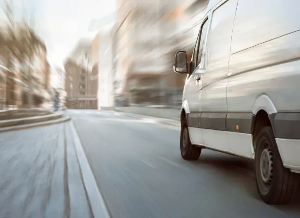 A white delivery van on an urban street. The background is blurred, suggesting the van is moving. 