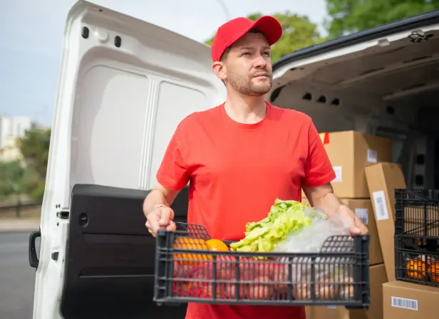 A delivery driver stands by the open door of his van, holding a plastic crate full of fruits and vegetables.