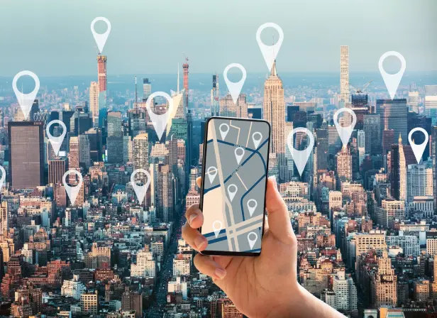 Aerial photograph of Manhattan with an overlay showing map pins at major locations. In the foreground a hand is holding up a mobile phone, with a screen showing a stylized map with pins.