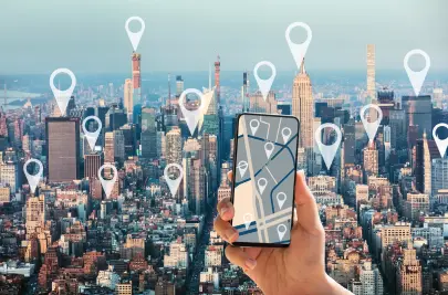 Aerial photograph of Manhattan with an overlay showing map pins at major locations. In the foreground a hand is holding up a mobile phone, with a screen showing a stylized map with pins.