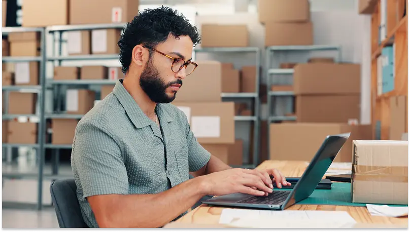 A bearded young man works at a computer in a warehouse setting with shelves full of boxes ready for delivery.