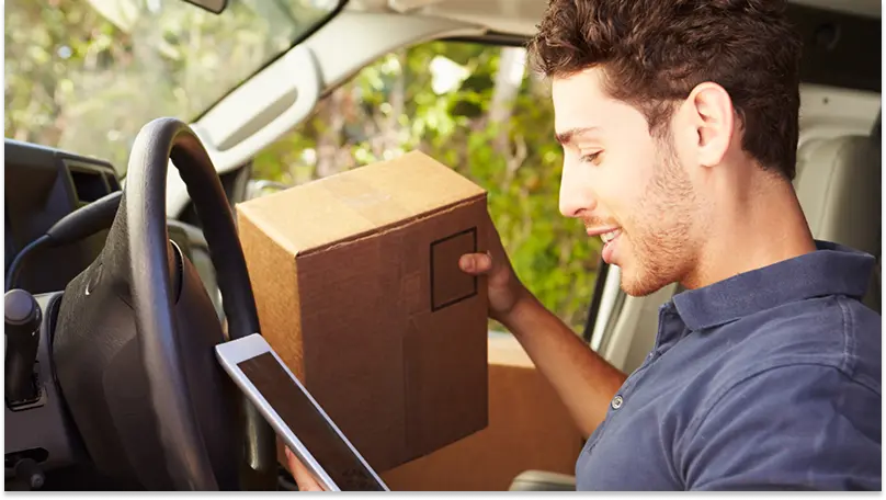 A delivery driver checks a tablet in his van while holding a package ready for delivery.