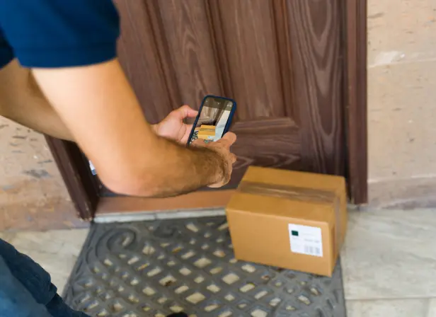 A delivery driver stands outside his vehicle holding a large cardboard box. He is wearing a mask and gloves.