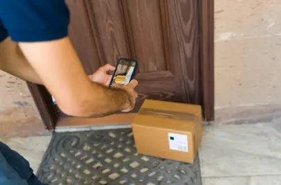 A delivery driver stands outside his vehicle holding a large cardboard box. He is wearing a mask and gloves.