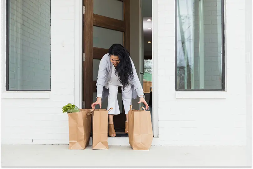 A woman collects three paper bags of groceries that have been dropped off on her doorstep
