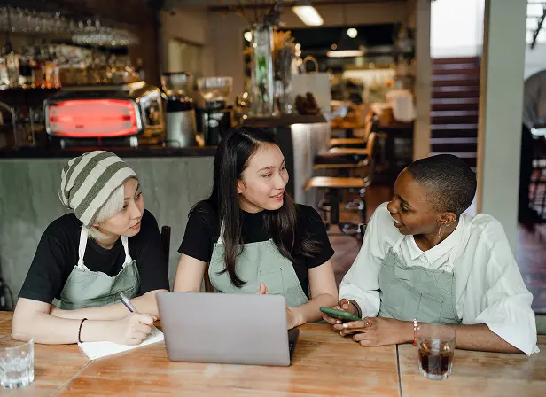 Three young women, wearing matching green aprons, sit at a restaurant table with an open laptop in front of them. They are deep in conversation and one is taking notes, possibly about their delivery logistics.