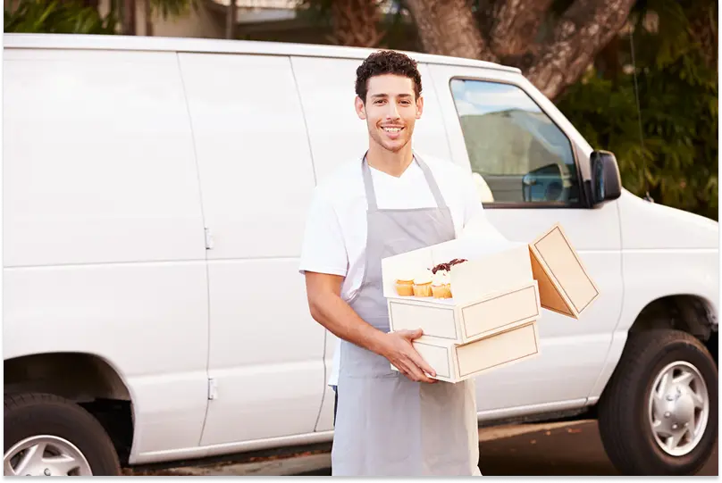 A man in an apron stands in front of a delivery van with a box of baked goods
