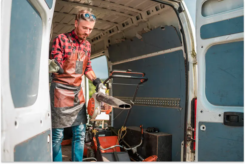 A young man wearing a leather apron and protective gloves unloads landscaping tools from a van.