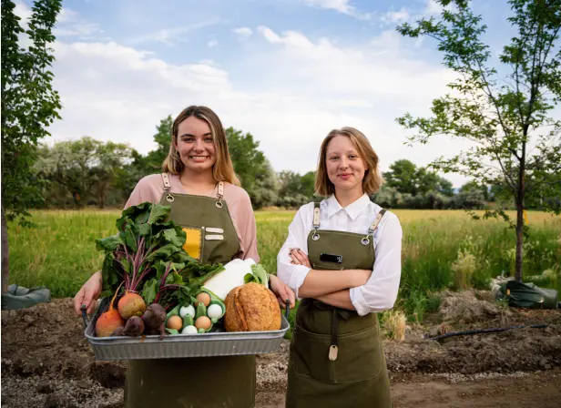 Two women stand in a field with a basket of fresh produce, eggs, and vegetables.