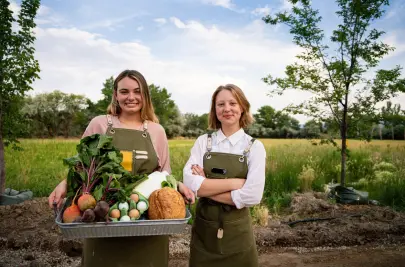 Two women stand in a field with a basket of fresh produce, eggs, and vegetables.