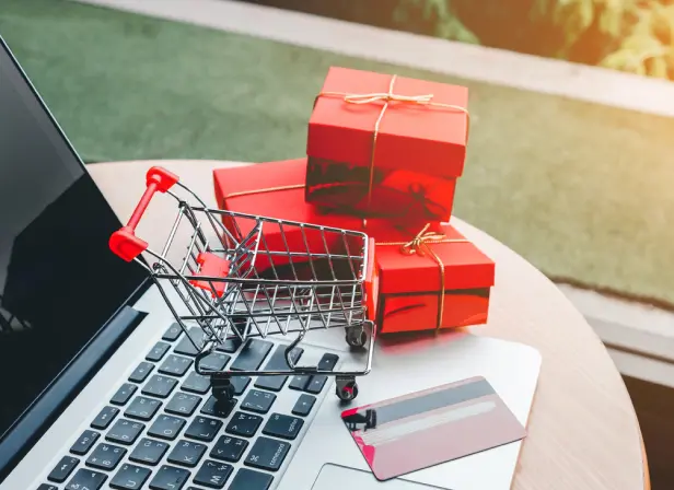 An open laptop on a patio table. On and around the laptop are a credit card, a miniature wire shopping care and a small pile of wrapped gifts.