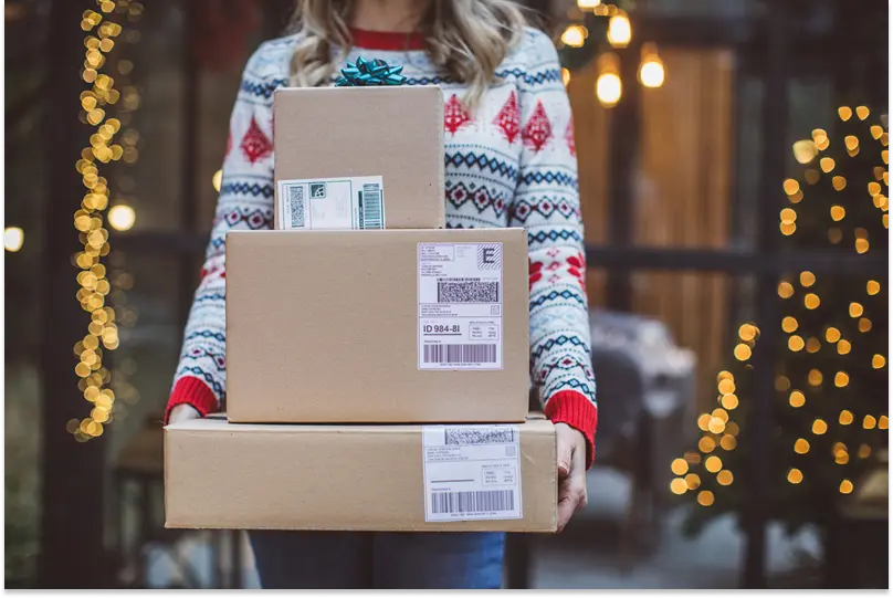 A woman in a holiday sweater holds three packages that have just been delivered.