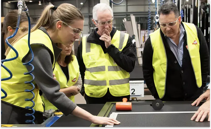 Four people in high-vis vests stand around a large table in a factory inspecting a window laid out flat.