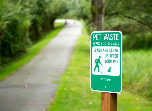 A along a scenic trail in a lush park reads “Pet waste transmits disease. Leash and clean up after your pet.”