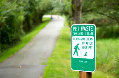 A along a scenic trail in a lush park reads “Pet waste transmits disease. Leash and clean up after your pet.”