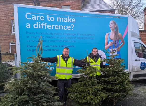 Pilgrims Hospice volunteers pose with Christmas trees in front of one of the charity’s trucks. The branding on the side of the truck says “Care to make a difference? Every fundraising event, challenge, fun-run, lottery or anything bought in our shops or donations help us to care for someone’s loved one. You really can make a difference, so get involved now!” The rest of the message is obscured by the volunteers and trees.