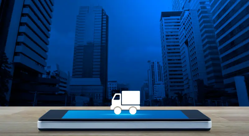 Composite image. An icon representing a delivery truck sits on a mobile phone that's lying horizontally on a table. In the background is a stylized image of tall city buildings.