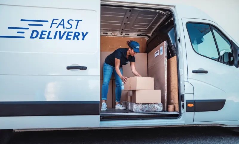 A white van with the words 'Fast Delivery' on the side. The sliding door is open, and a woman is busy sorting delivery packages inside.