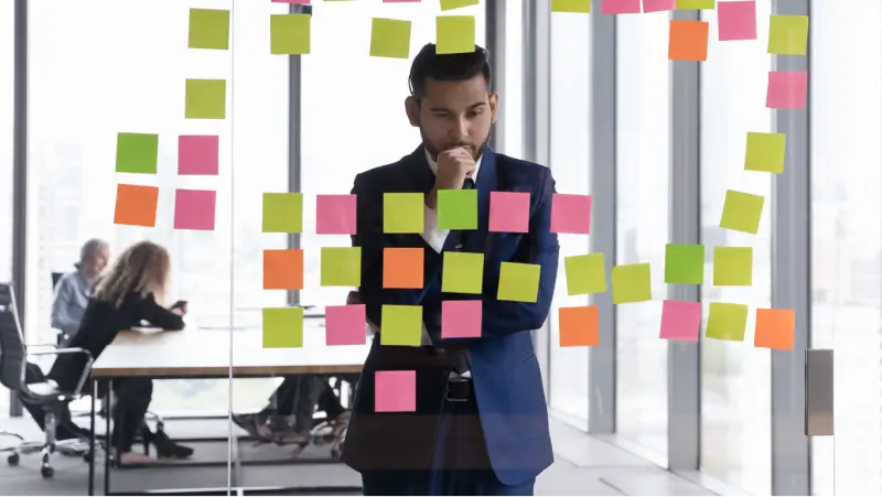 A man in a business suit stands in front of a glass wall full of brightly colored sticky notes. One arm is across his body and the other hand is held to his mouth, indicating he is deep in thought.