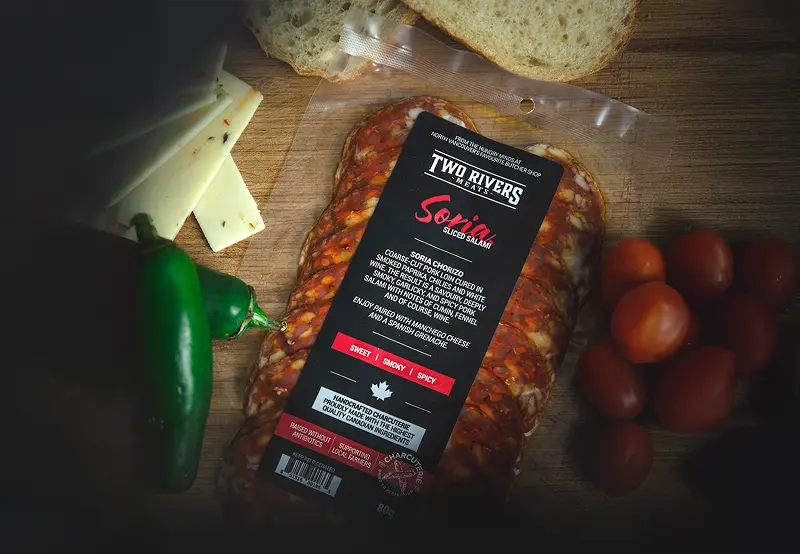 A pack of Two Rivers Meats salami staged on a wooden table with chili peppers, cheese, bread and tomatoes.