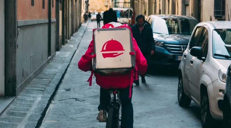 A cyclist wearing a food delivery backpack rides down a narrow Italian city street.